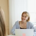 A young woman with a laptop smiling at another person in an indoor business setting.